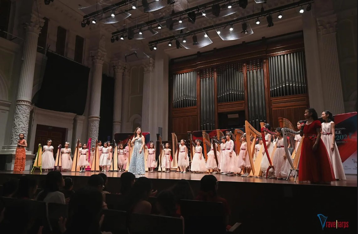 Katryna leading a group harp performance on stage, demonstrating harp technique with students during a concert recital